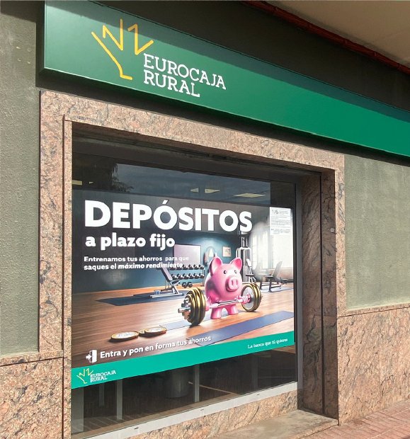 Fachada de oficina de Eurocaja Rural con el escaparate vinilado con el cartel de la campaña; logo de Eurocaja Rural en banderola superior verde