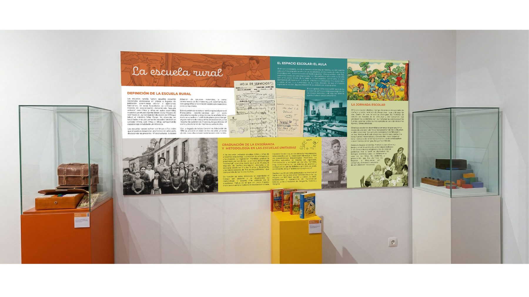Sala expositiva del museo con el panel «La escuela rural» instalado en pared, dos vitrinas con maletines de cuero a izquierda y caja de regletas a derecha, y peana naranja con libros entre ambas