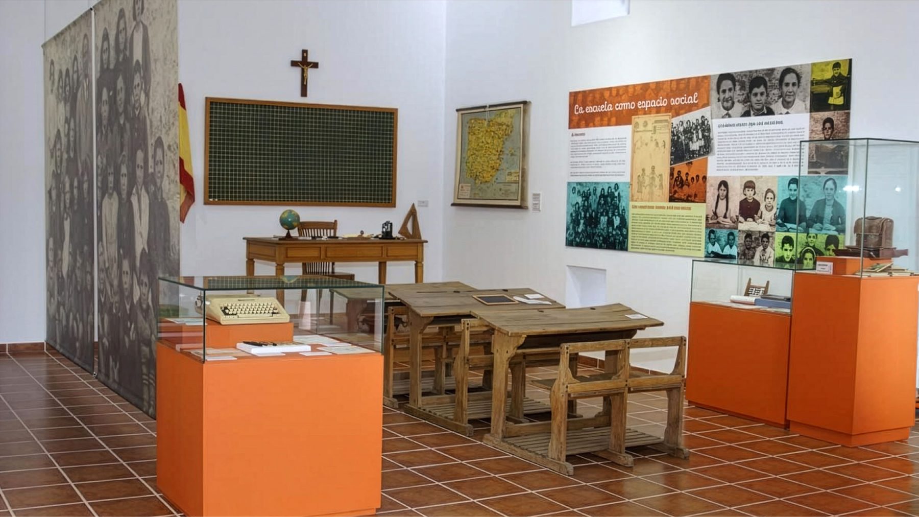 Otra sala del museo con foto histórica de aula reproducida a tamaño mural en la pared del fondo, pizarra escolar, mesa del maestro con globo terráqueo, pupitres de madera originales, banderas, mapa regional y crucifijo