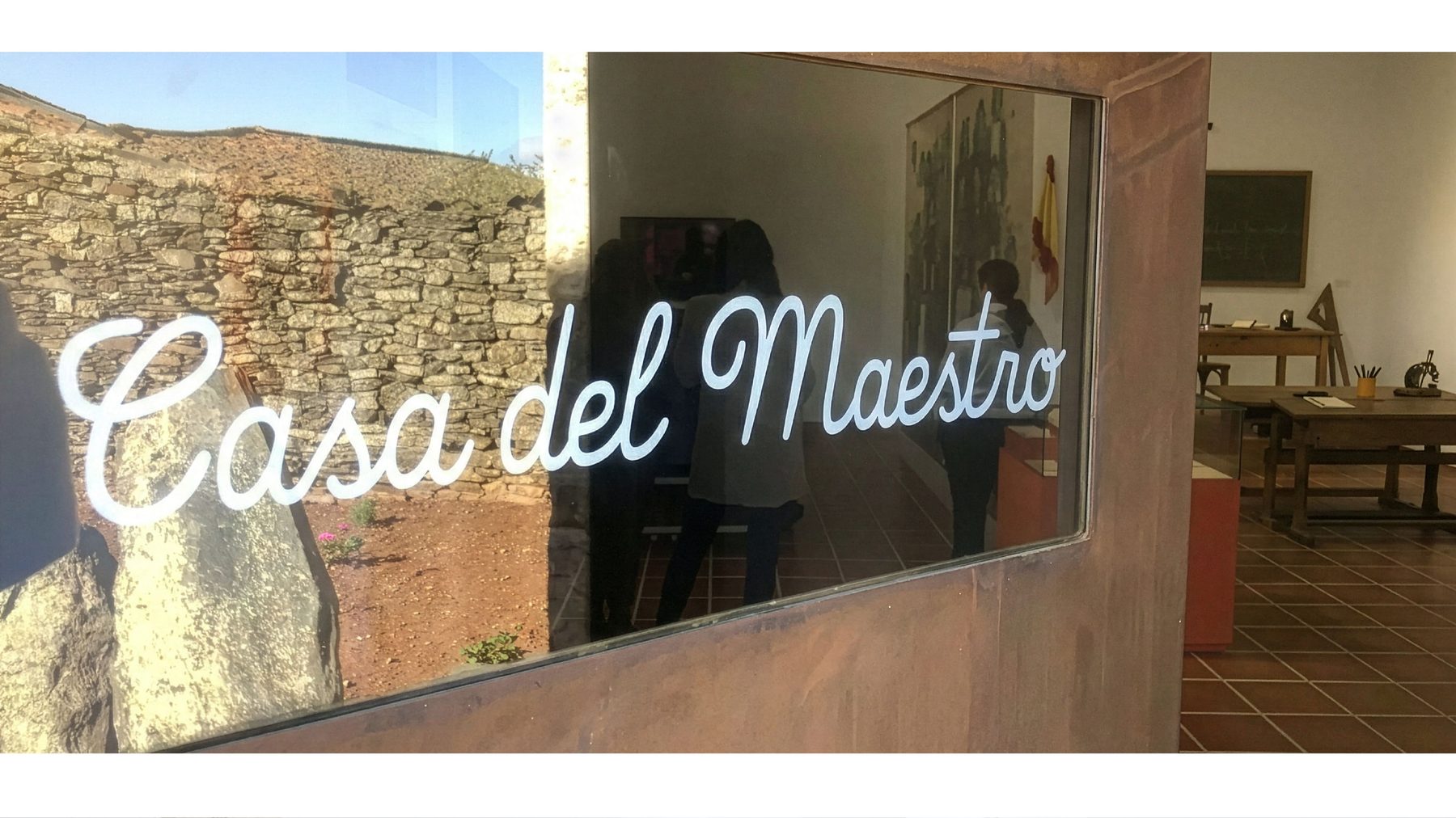 Rótulo principal del museo en vinilo blanco aplicado a una mampara de cristal con marco de acero corten oxidado, con el letrero «Casa del Maestro» en cursiva manuscrita y visitantes reales reflejándose en el cristal