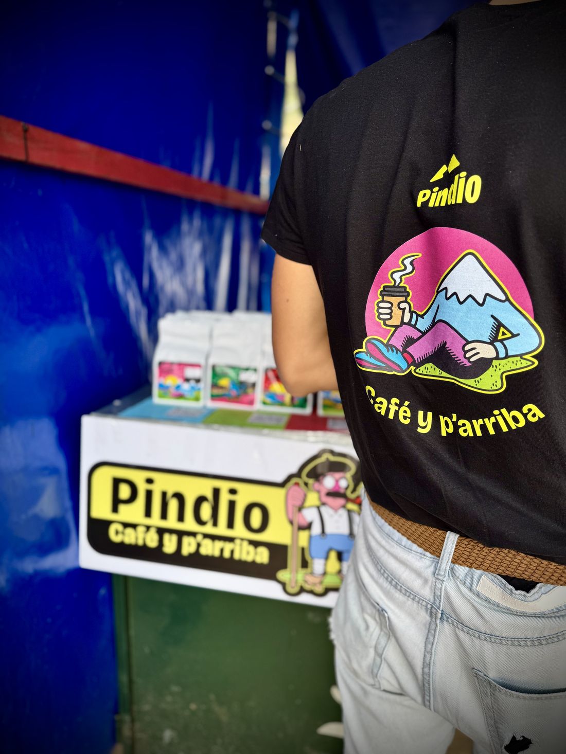 Foto real: empleado de cafetería con la camiseta Pindio y display de bolsas y cartelería al fondo