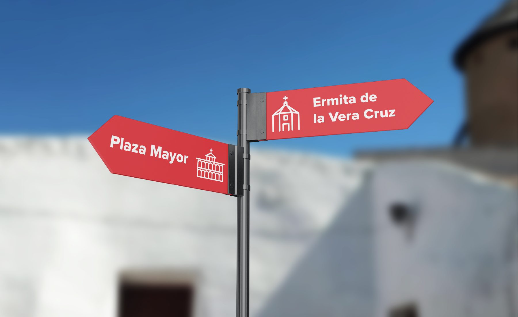 Señales direccionales rojas montadas en farola: «Plaza Mayor» con flecha izquierda y «Ermita de la Vera Cruz» con flecha derecha, sobre el muro encalado de una calle del pueblo