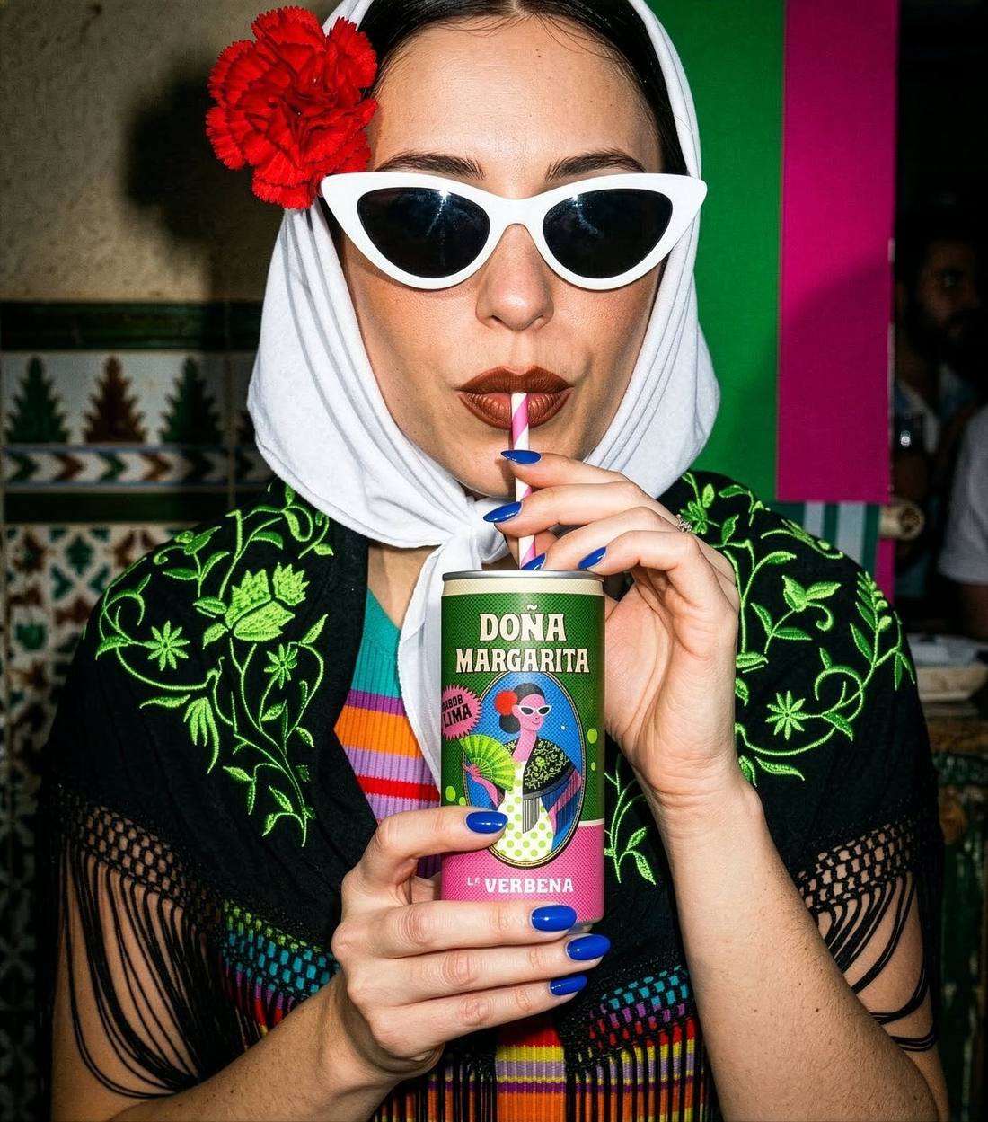 Mujer joven con pañuelo blanco, gafas blancas y clavel rojo en la cabeza, vestida con mantón verde bordado, bebe Doña Margarita con pajita rosa y blanca